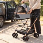 Hand lifting the gray carrier with a tabby cat inside straight up off the stroller frame beside an open car door