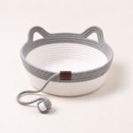 snugweave-rope-cat-basket-gray Gray and white rope cat basket with cat-ear handles and a knotted gray rope ball, empty, on a cream backdrop