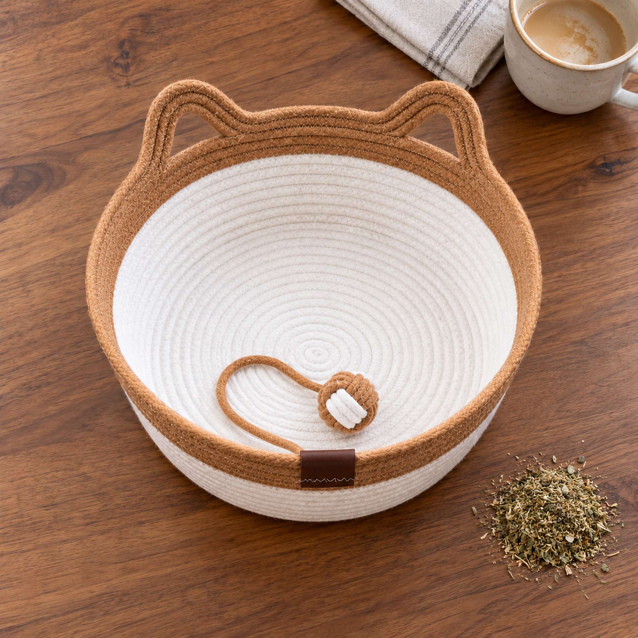 Flat lay on a dining table: a caramel rope cat basket bed with the rope ball, loose dried catnip and a coffee mug